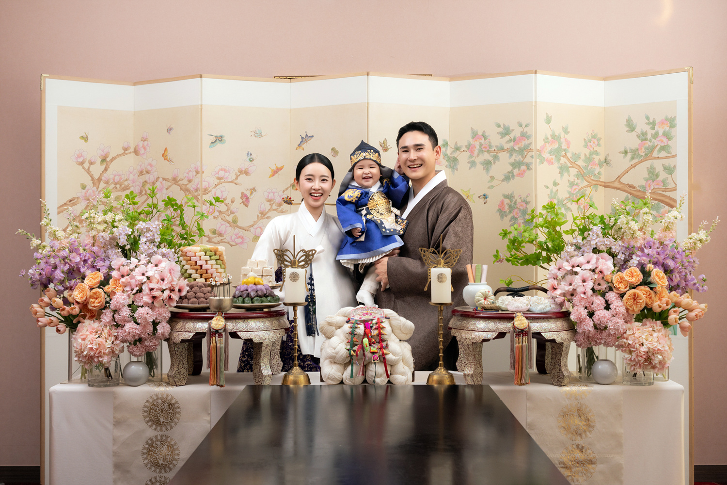 A Korean baby in soft hanbok held close by family, the quiet register of a hundred-day baek-il celebration at home.