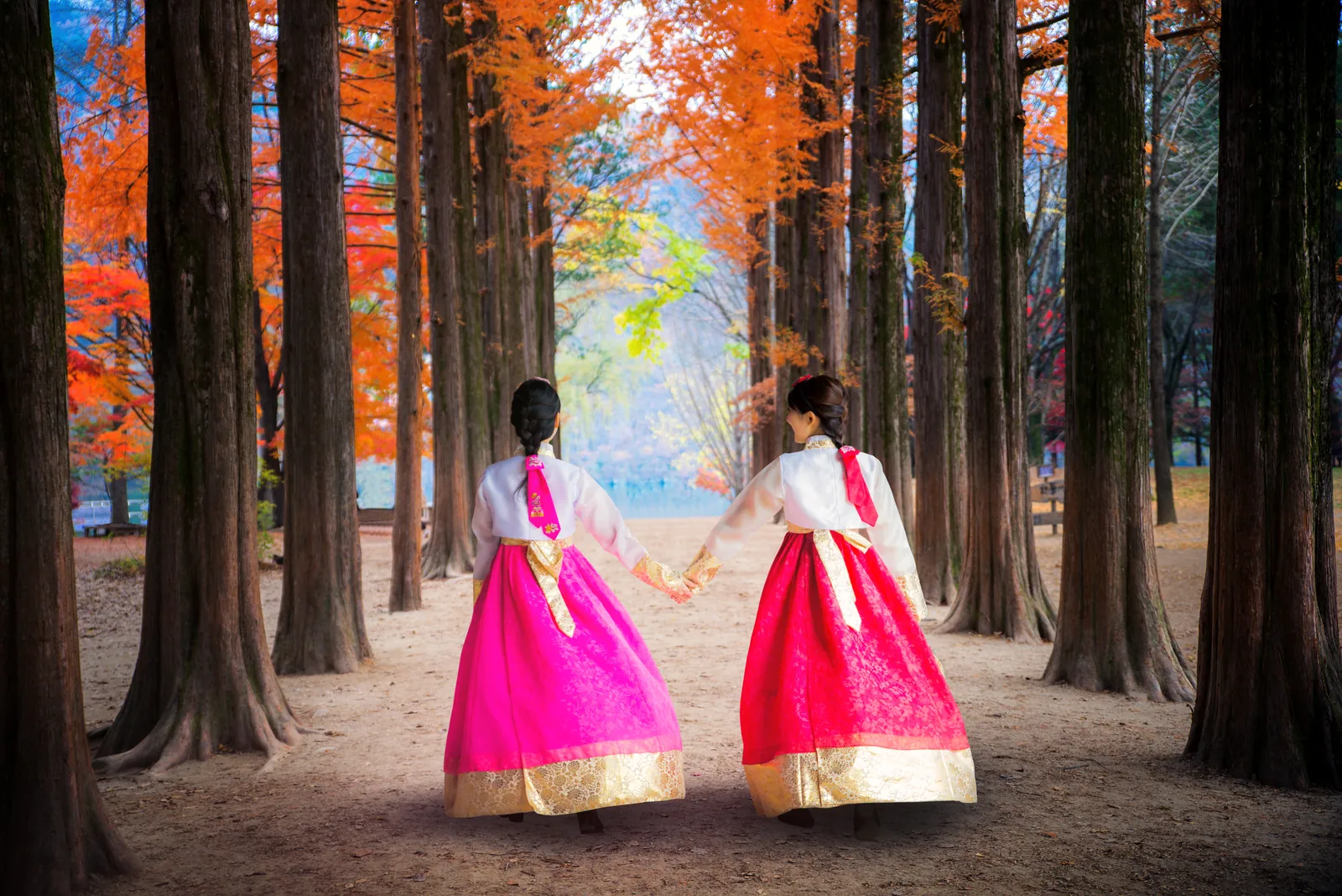 Two women in bright hanbok walking hand in hand through an autumn forest
