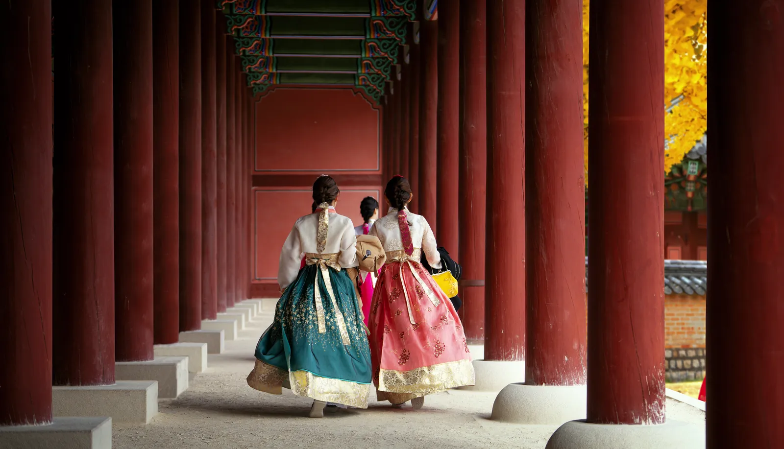 Two women in hanbok walking along a red colonnade in autumn